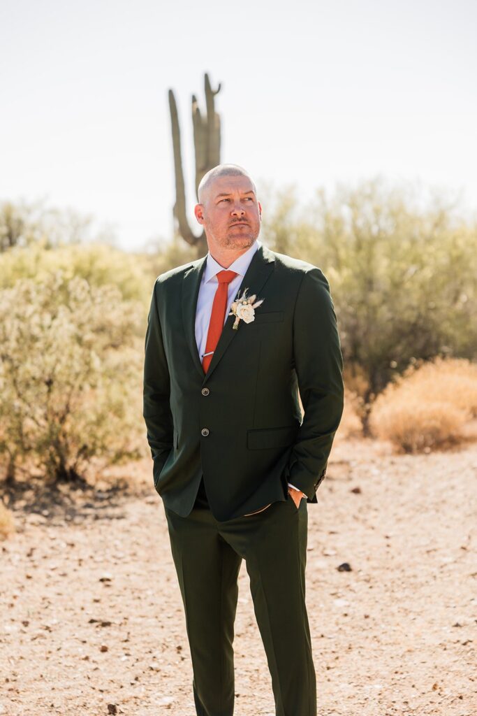 Groom in a forest green suit and rust tie standing in the superstition paseo desert with cactus in the background.