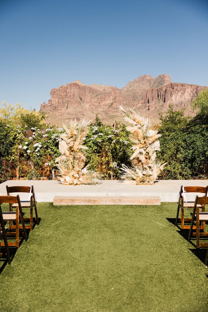 Ceremony site at The Paseo featuring pampas grass floral pillars, wooden chairs, and the Superstition Mountains backdrop.