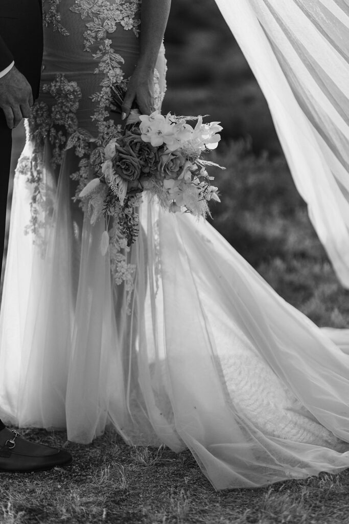 Black and white photo of bride holding a cascading boho bouquet, showing lace train details of her wedding dress.